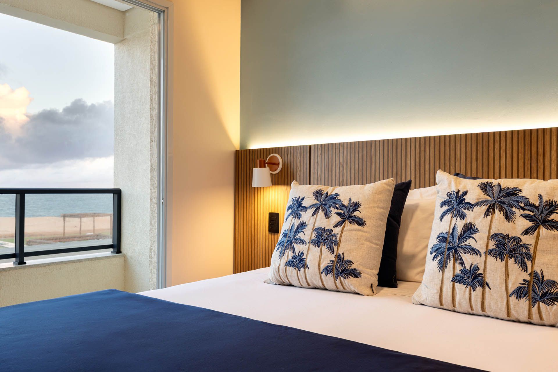 Quarto de hotel moderno com almofadas e cama em tons de azul e branco com estampa de palmeiras, e vista para o mar através de uma grande janela.