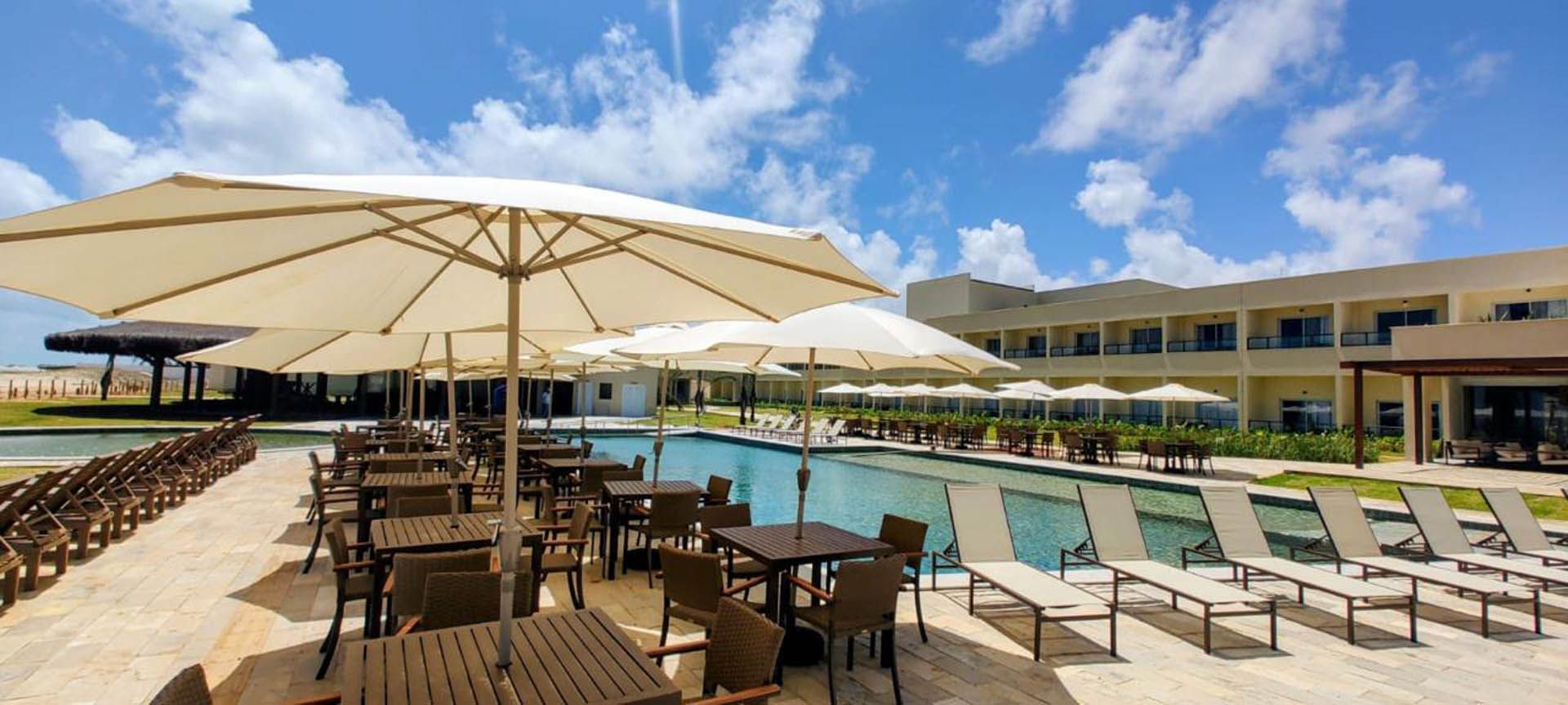 Piscina ensolarada de resort com guarda-sóis brancos, espreguiçadeiras e água azul sob um céu parcialmente nublado.