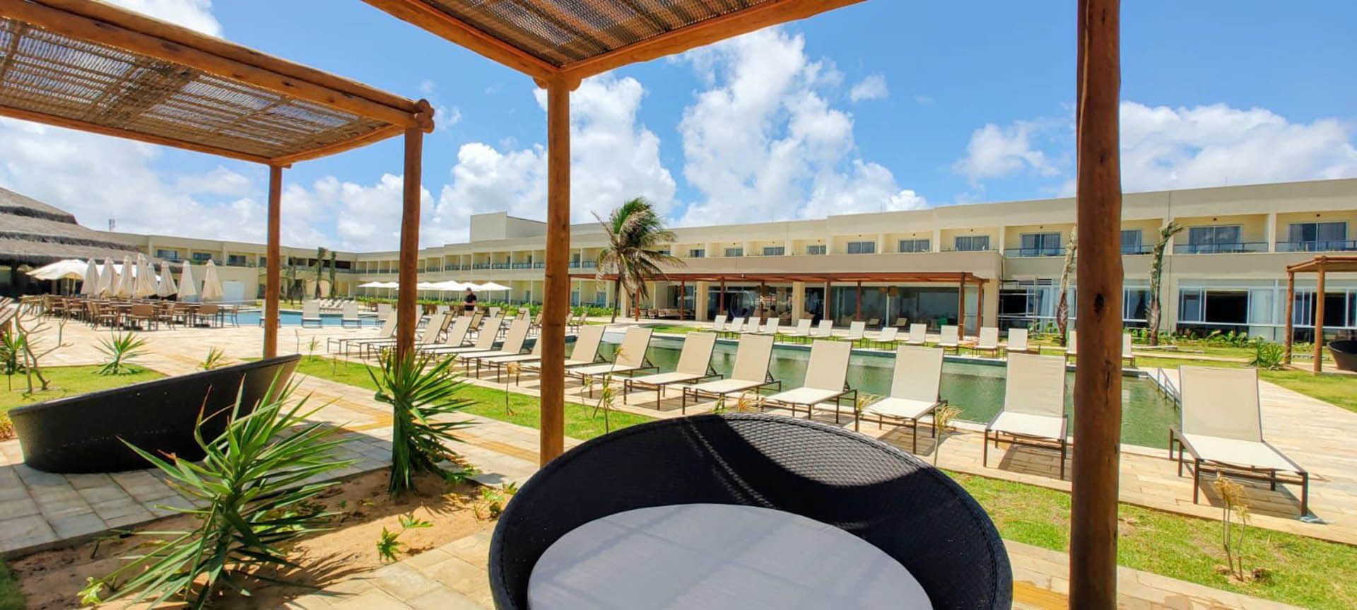 Área da piscina do hotel com água turquesa, espreguiçadeiras, guarda-sóis brancos e um deck de azulejos.