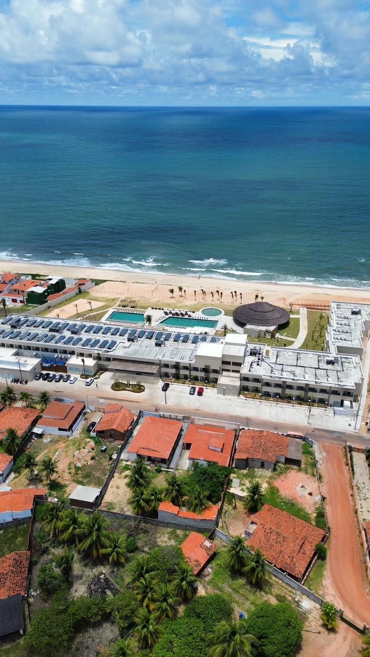 Vista aérea de um resort de praia com oceano turquesa, areia branca e edifícios de telhados vermelhos em primeiro plano.