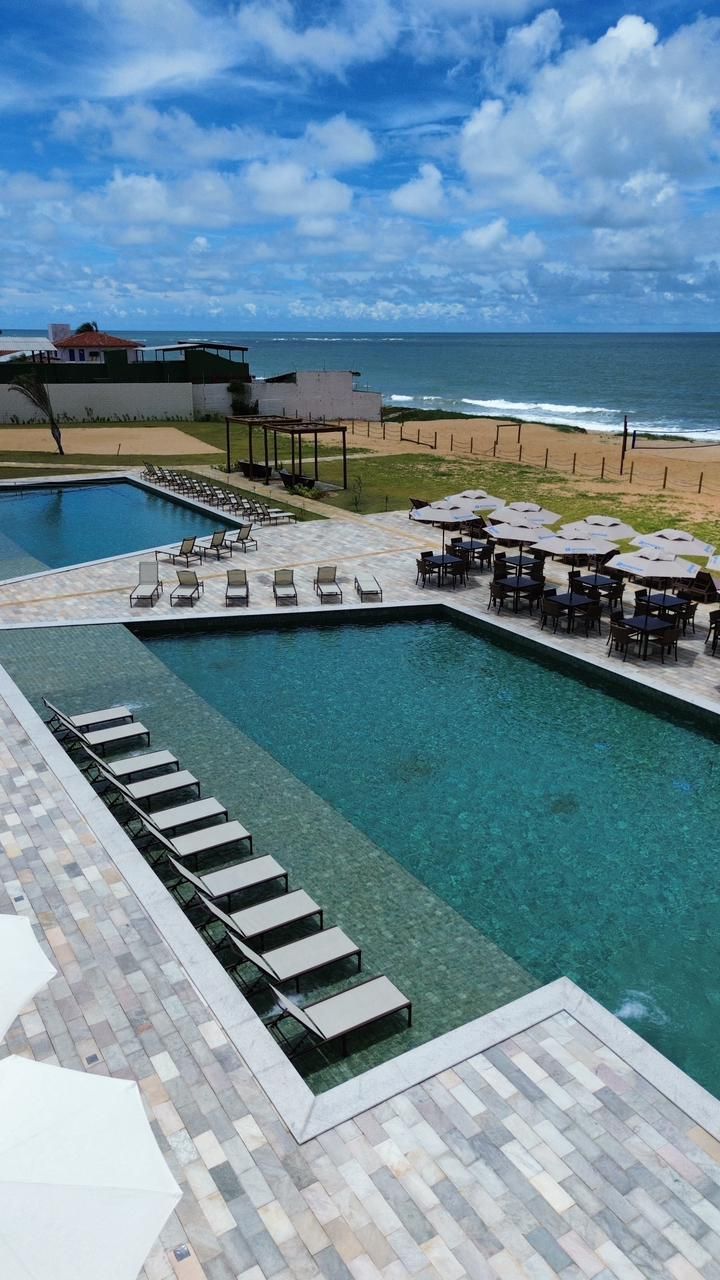 Piscina de resort à beira-mar com espreguiçadeiras, guarda-sóis e uma praia sob um céu azul parcialmente nublado.