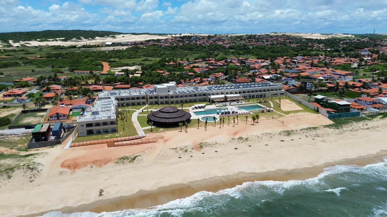 Vista aérea de um resort à beira-mar com piscinas, areia e uma cidade costeira ao fundo.