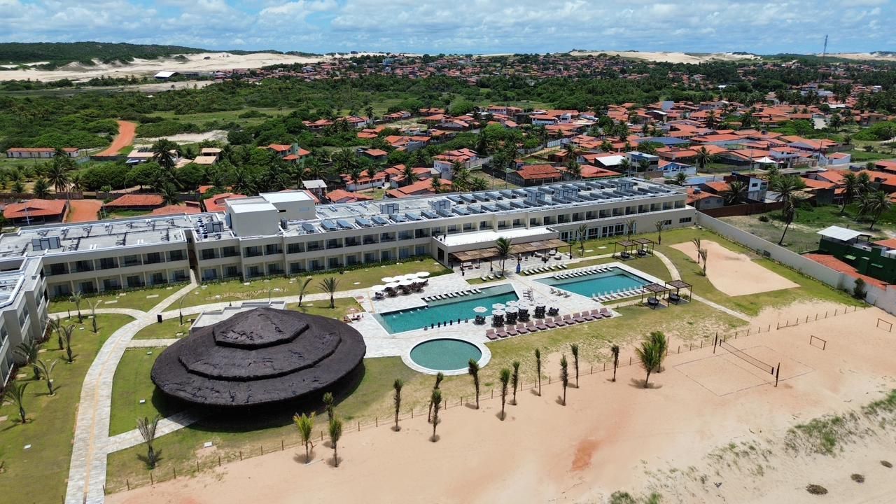 Vista aérea de um resort com piscina, pavilhão circular com teto de palha e pátio à beira-mar, próximo a uma cidade litorânea.