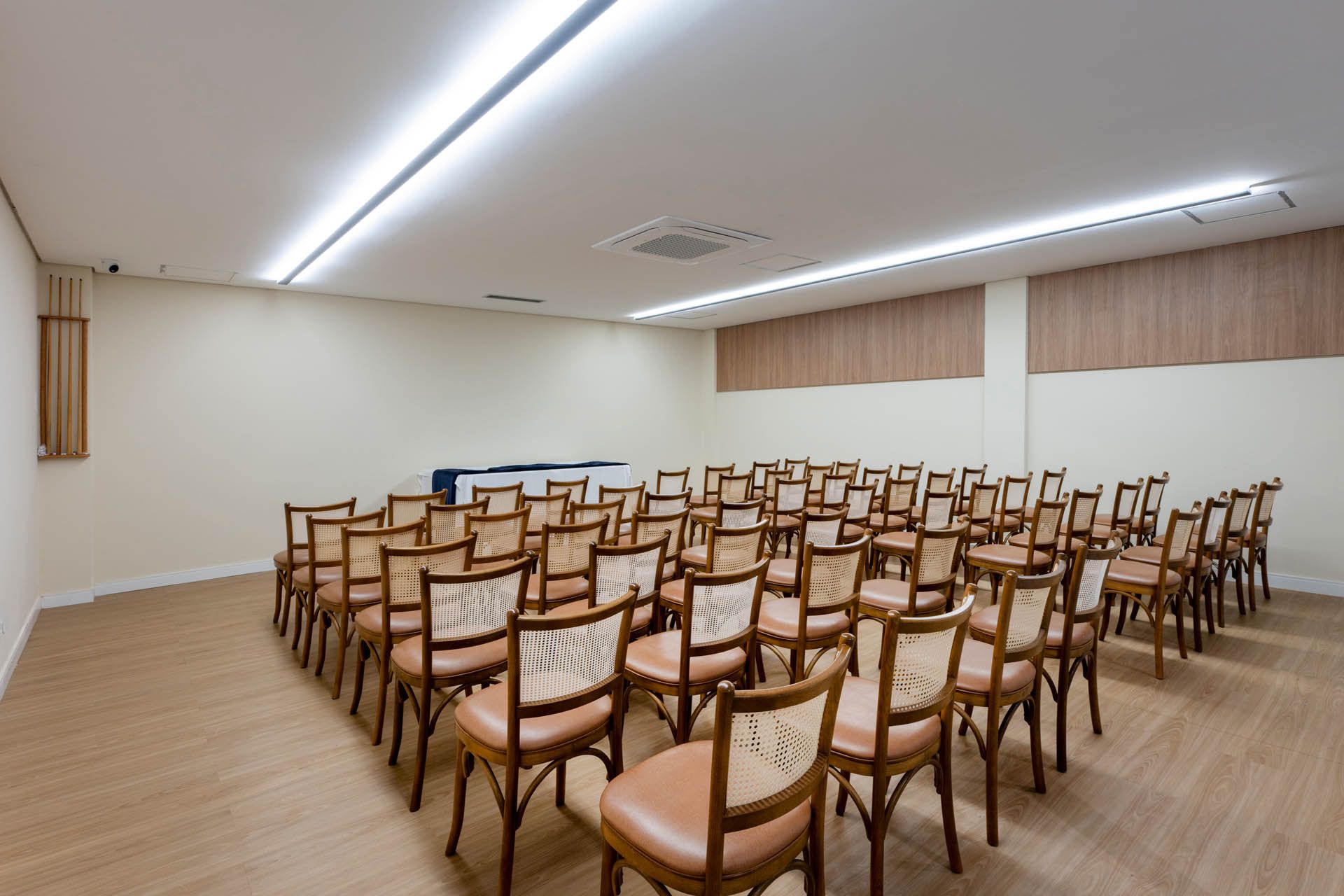 Sala de conferências vazia com fileiras de cadeiras de madeira e teto branco com iluminação linear.