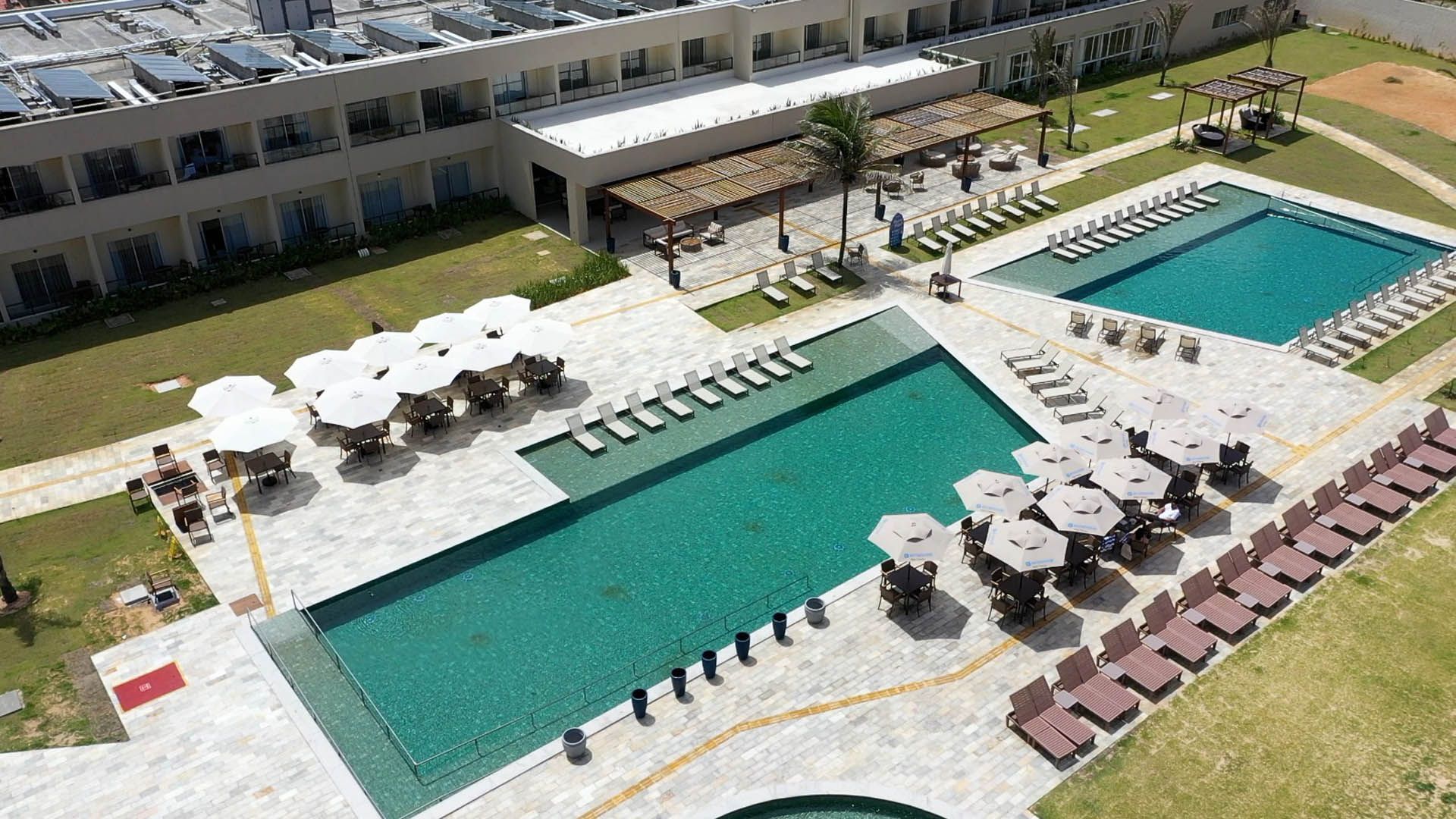 Vista aérea de duas piscinas turquesa com espreguiçadeiras e guarda-sóis no pátio de um resort.