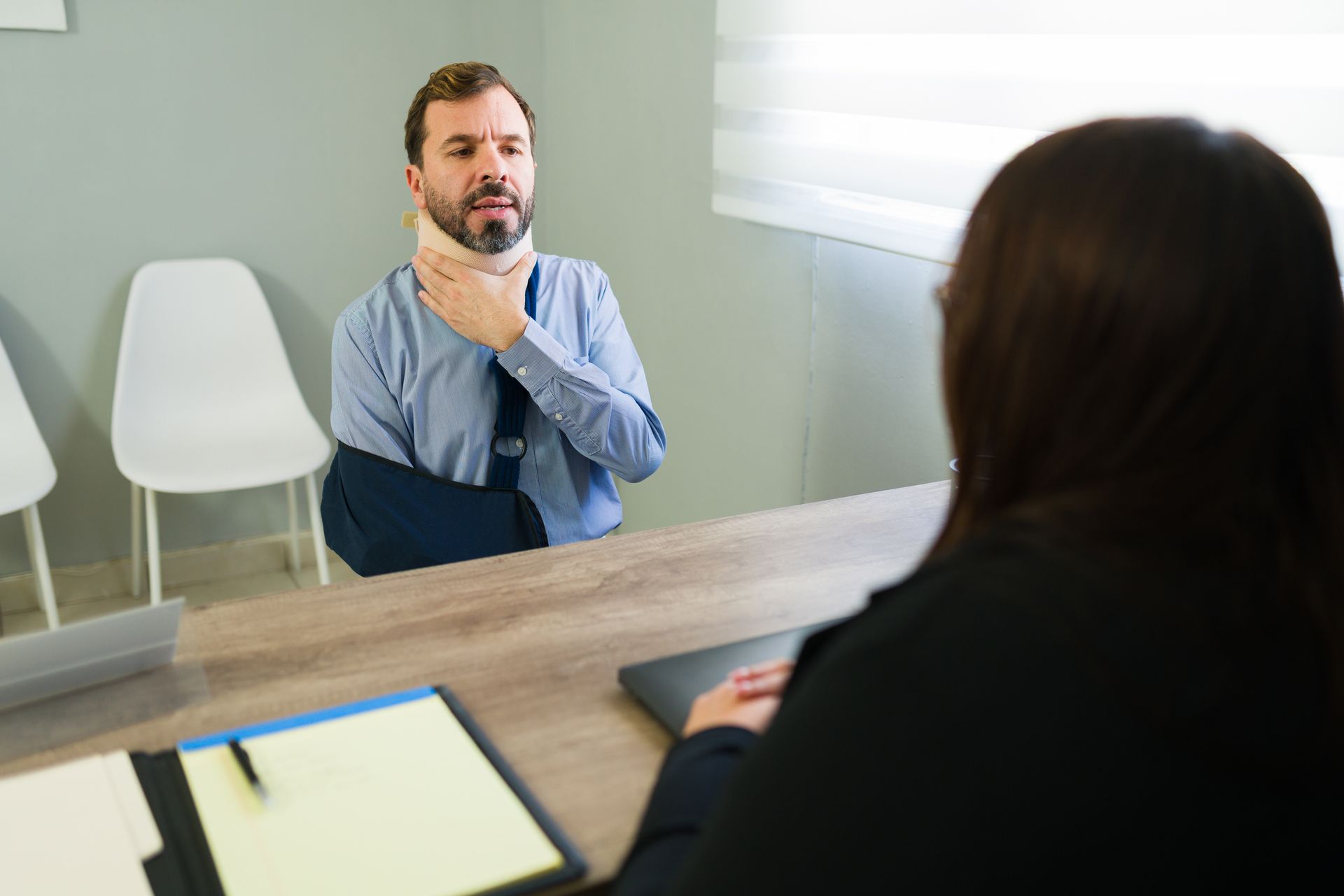 A professional in a neck brace and arm sling sits at a desk, talking to someone across from them in an office.