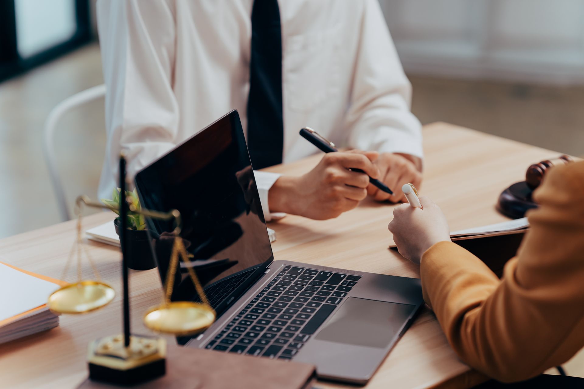 Two professionals sit at a desk with a laptop and scales of justice, discussing a document during a meeting.
