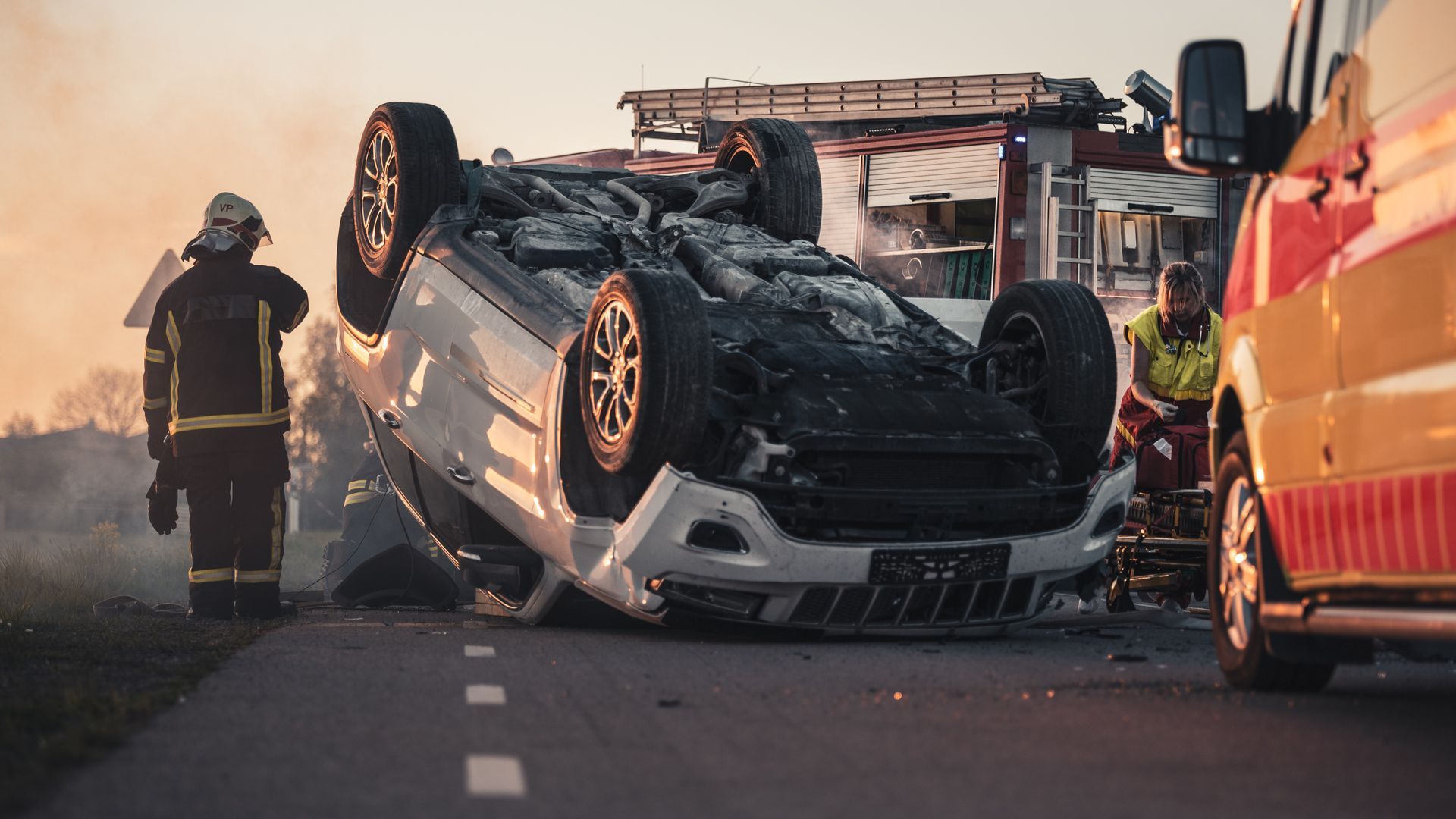 A white car flipped on its roof at a crash scene, with a firefighter standing nearby and an ambulance parked in view.