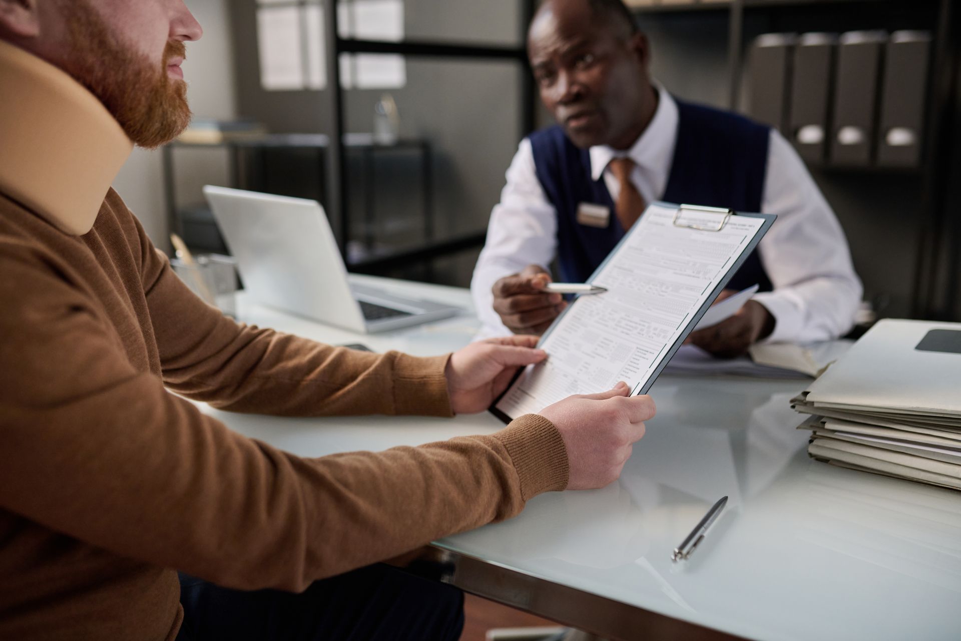 A person wearing a neck brace reviews a document with an office worker at a desk.