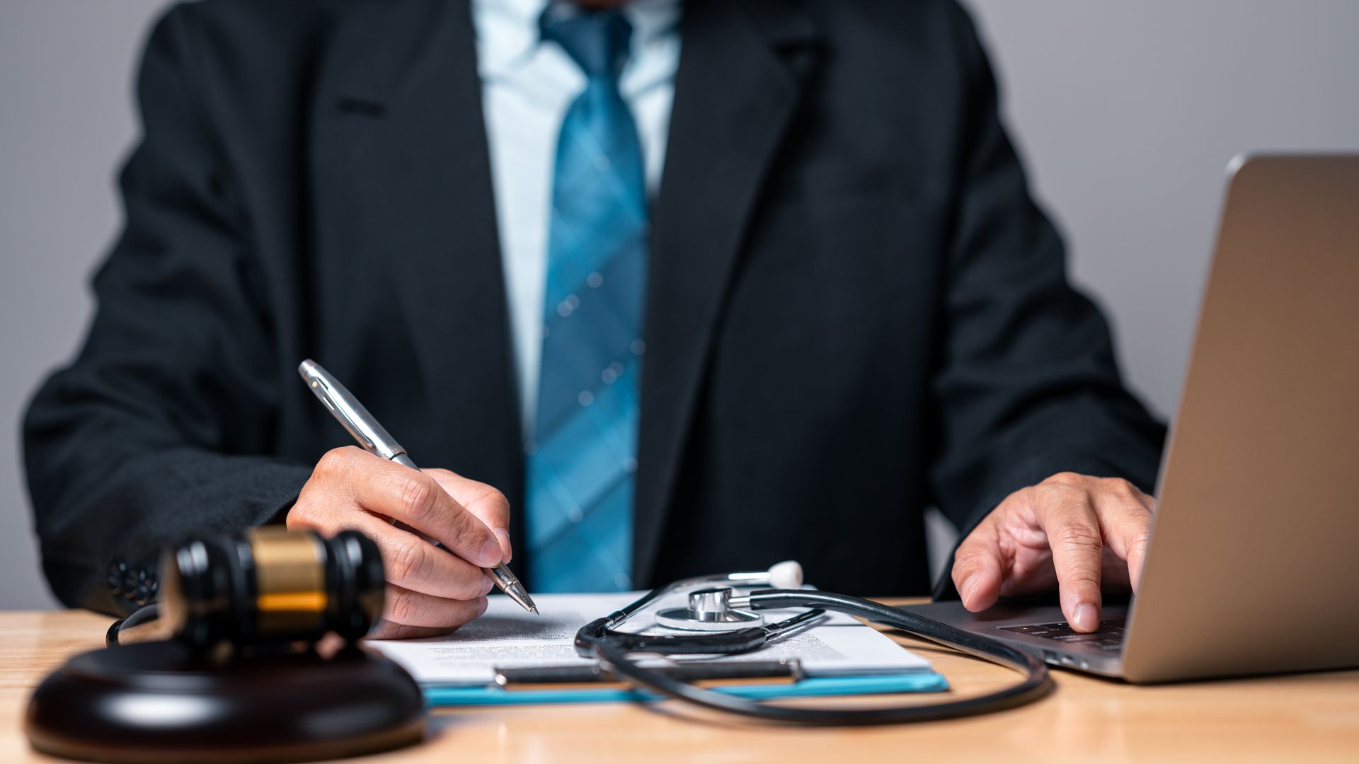 A person in a suit writes on a document next to a gavel, stethoscope, and laptop on a desk. A person in a suit writes on a document next to a gavel, stethoscope, and laptop on a desk.