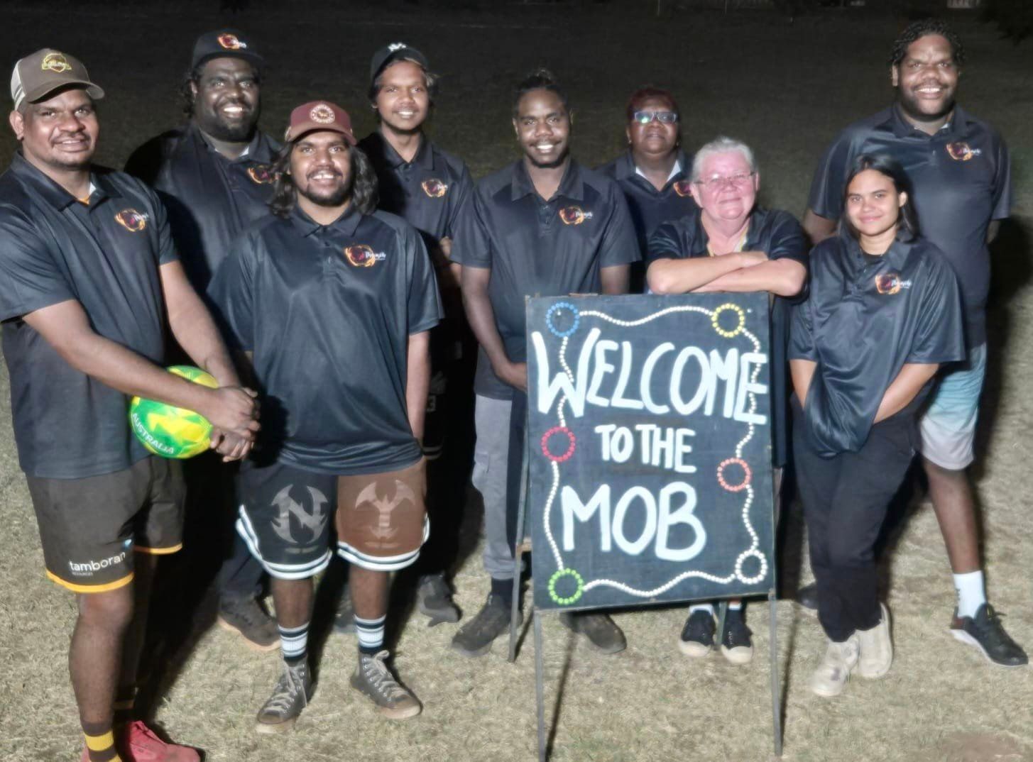 A group of people, mostly men, in dark shirts stand together, some smiling. They hold a sign that says 