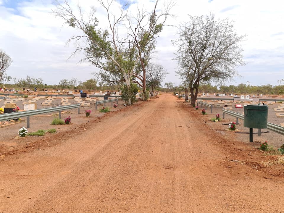 Dirt road through a cemetery with rows of graves. Trees line the road under a cloudy sky.