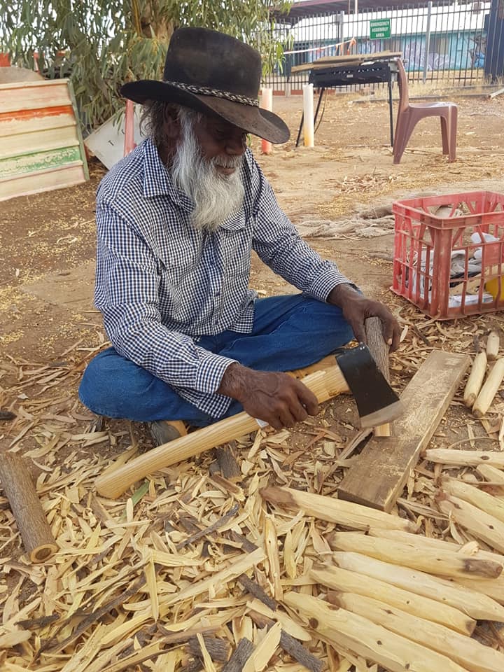 Man with a long white beard and hat, sitting on the ground and using an ax to carve wood; outdoors.