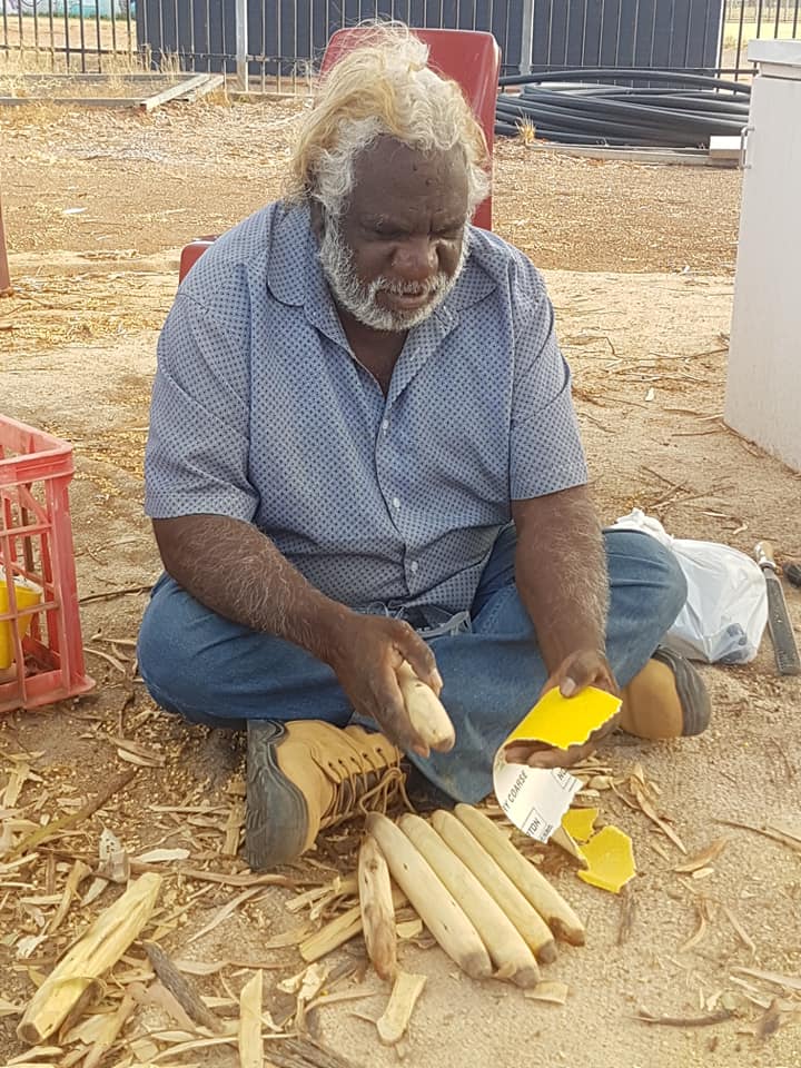 A man with white hair carves wood while sitting on the ground outdoors. He wears blue jeans, a button-up shirt, and boots.