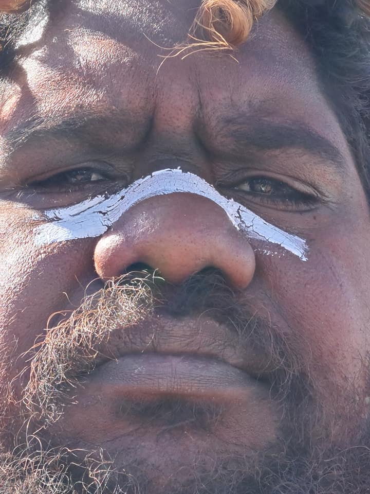 Close-up of a man with dark skin, a white line of paint across his nose, and a serious expression.