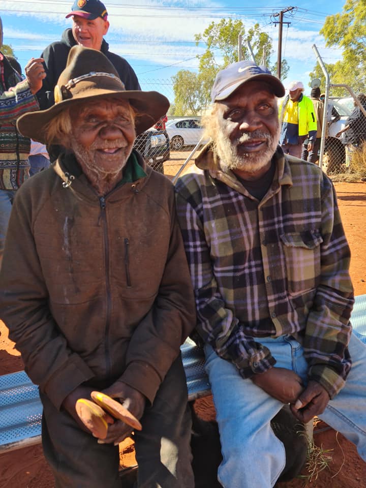 Two older Indigenous men smiling, seated outdoors, one in a hat and jacket, the other in a plaid shirt and cap; dirt ground.