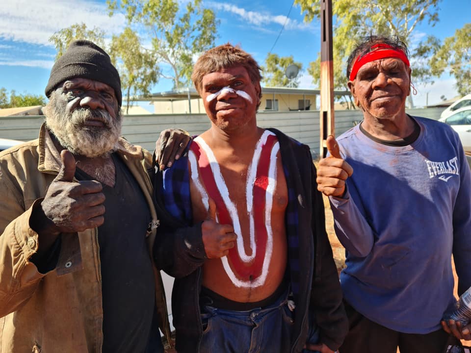 Three Indigenous men giving thumbs up; one with face and chest paint, another with a red headband. They are outdoors.