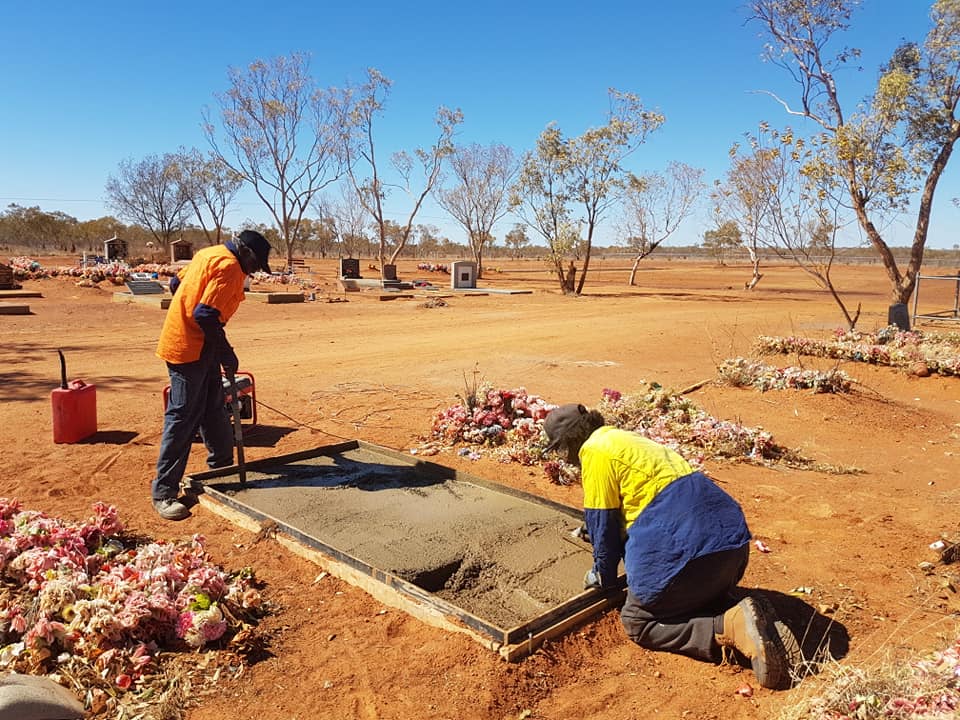 Two workers pour concrete on a grave in a sunny, dry cemetery. One stands, the other kneels, both in work clothes.
