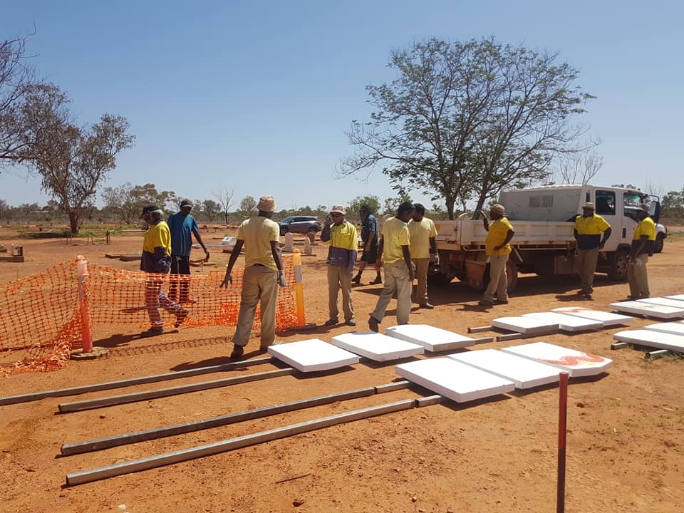 Workers in yellow shirts assemble solar panels in a sunny, arid outdoor setting, with a truck in the background.