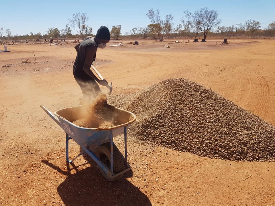 A person shovels gravel from a pile into a wheelbarrow in a dry, dusty, rural setting.