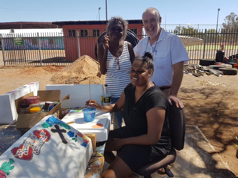 Three people, two Indigenous women and a man, smile for the camera while painting outdoors.