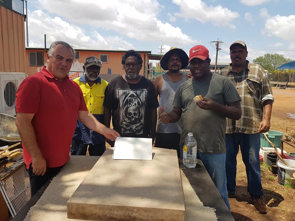 A group of six men standing outside, some holding a document. The man in red points to the document on a stone surface.