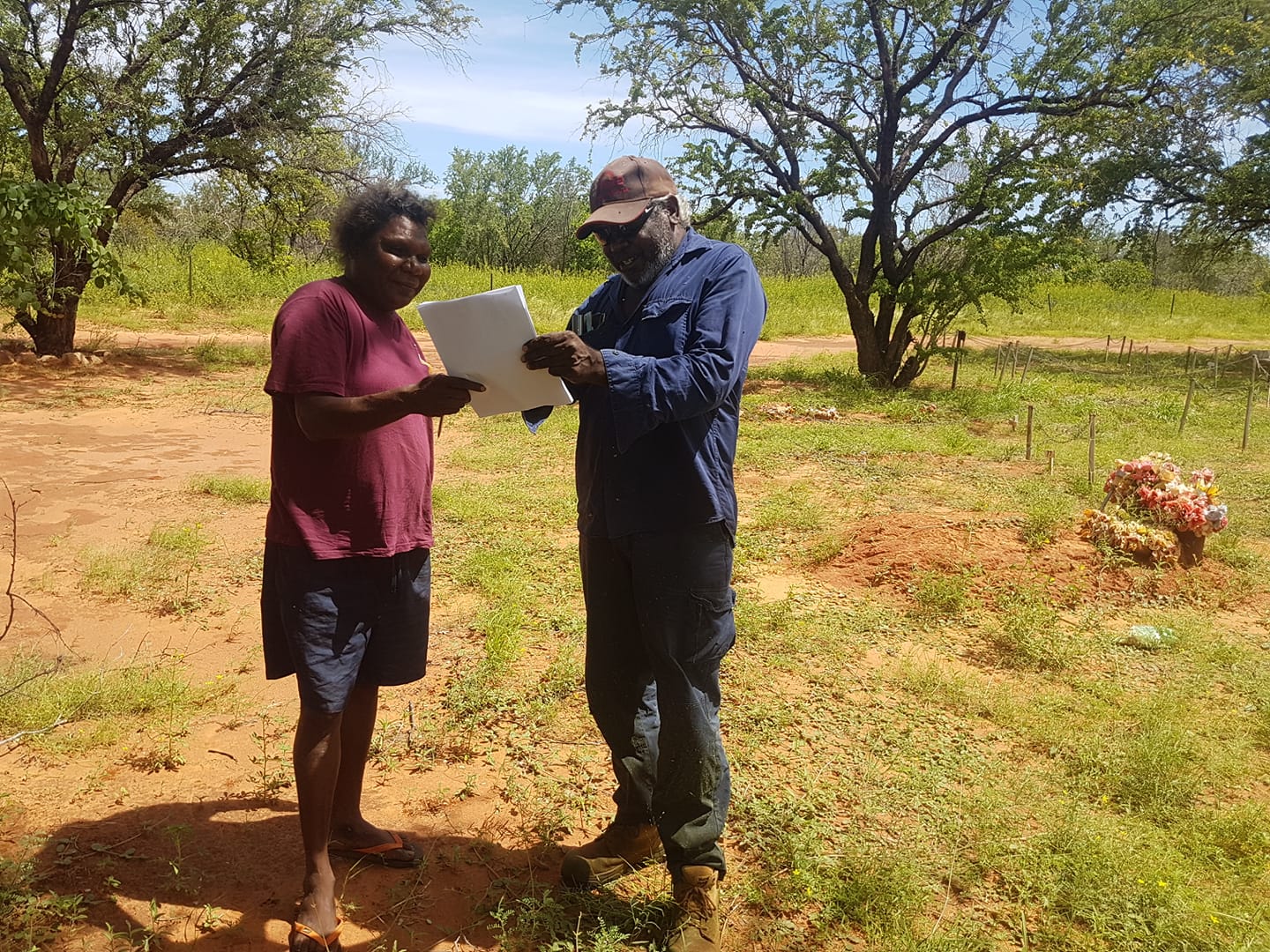 Two people, one in a red shirt, the other in a blue shirt and hat, review papers outdoors on dry, grassy land.