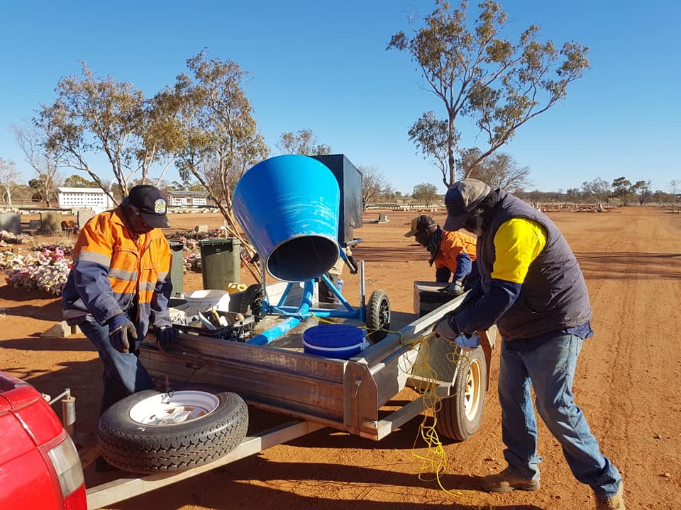 Three workers in safety vests prepare a blue cement mixer on a trailer in a sunny, outdoor setting with dry, reddish earth.
