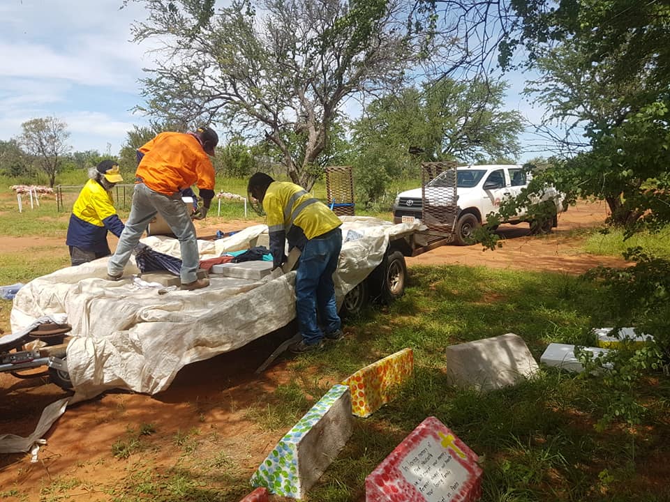 Three people in work vests and a truck unload items from a trailer in a grassy, outdoor area. Scattered debris is visible.