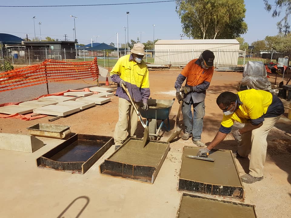 Three construction workers pouring and smoothing cement in square molds outdoors. They wear work clothes and safety gear.