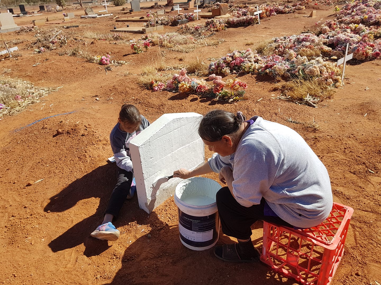 Two people paint a white headstone in a cemetery.