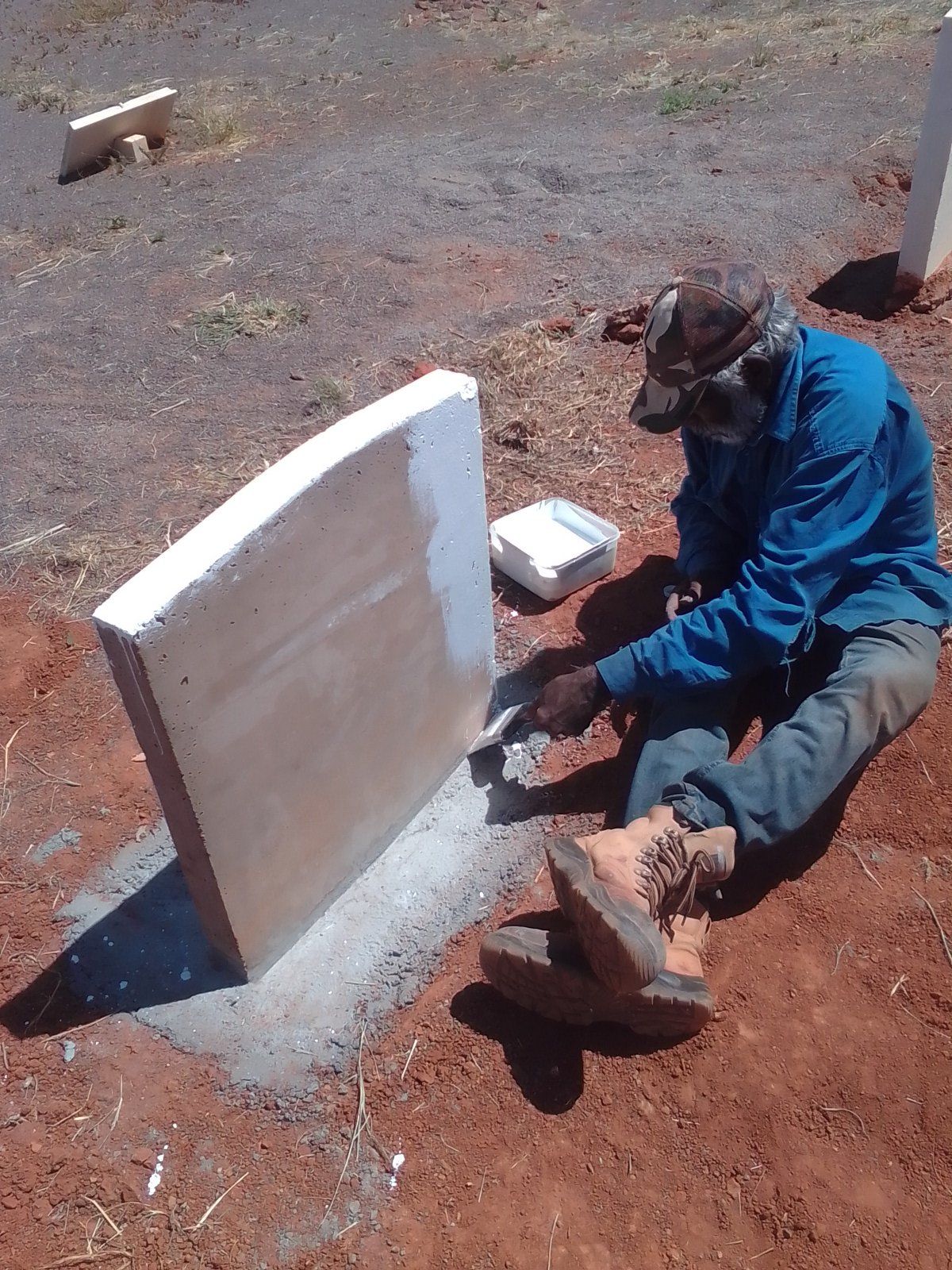 Man painting a white edge on a concrete post outdoors on a red dirt surface. He wears a blue shirt, hat, and boots.