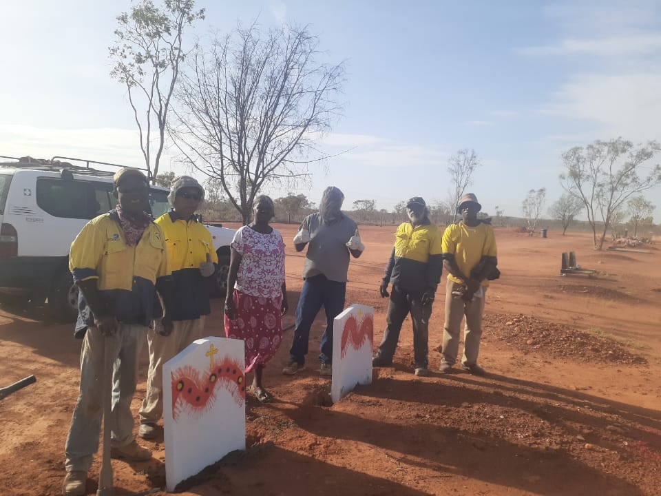 A group of people stand near two markers in a red dirt field. They are likely working on a project, in a rural setting.