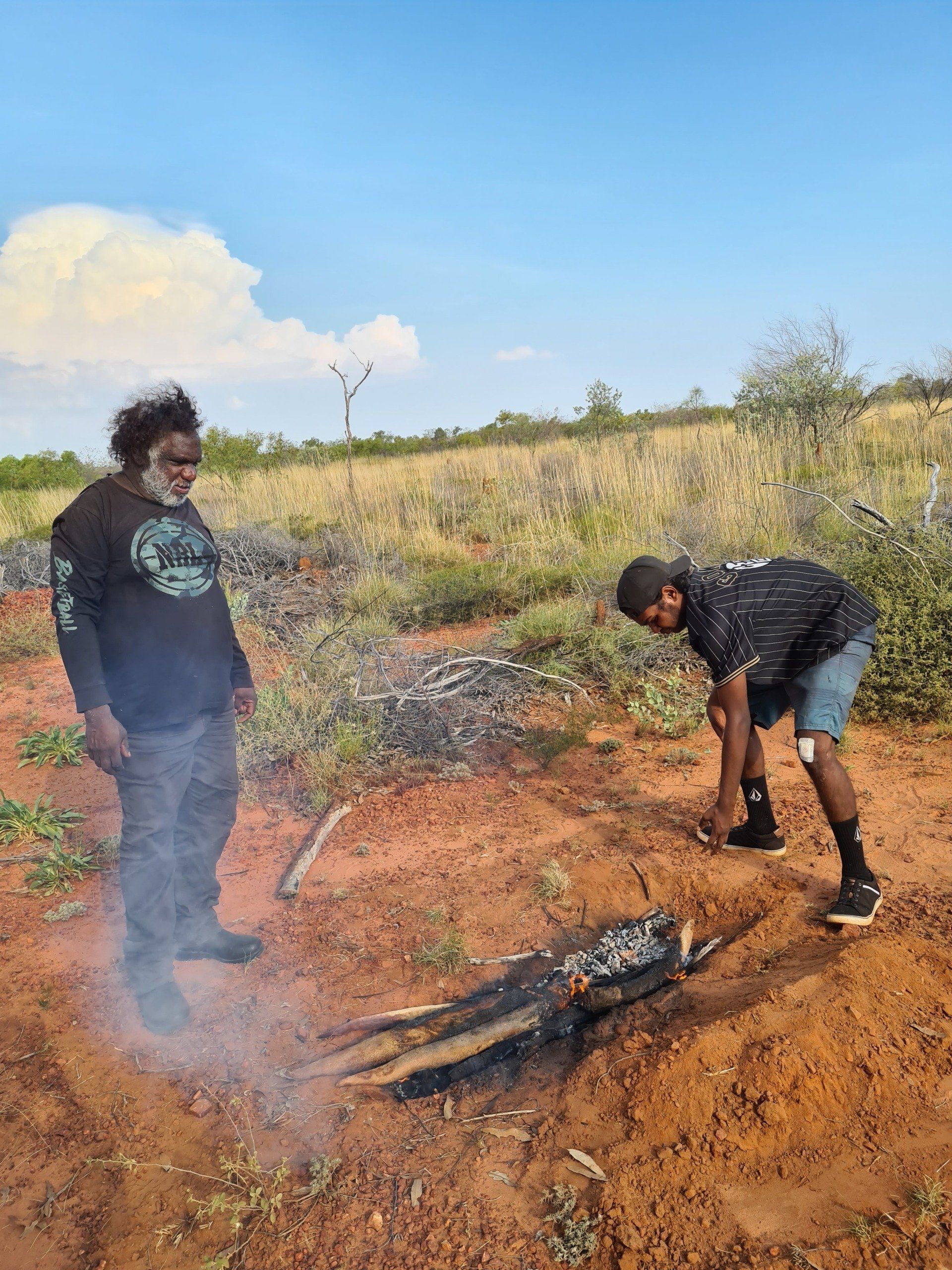 Two Indigenous men near a smoldering log on red earth under a blue sky.