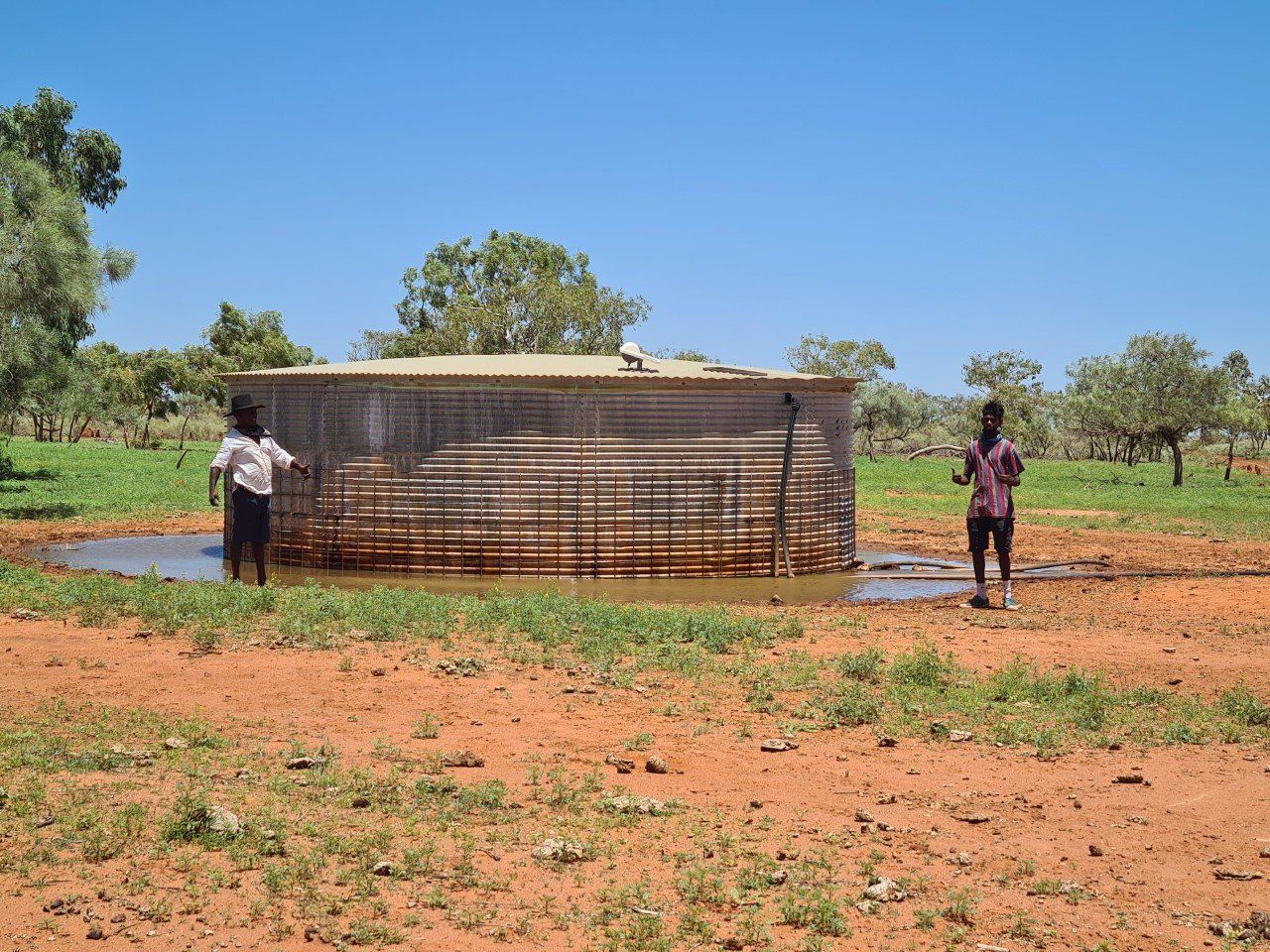 Two people standing near a metal water tank in a field of red dirt and grass; trees and blue sky in the background.