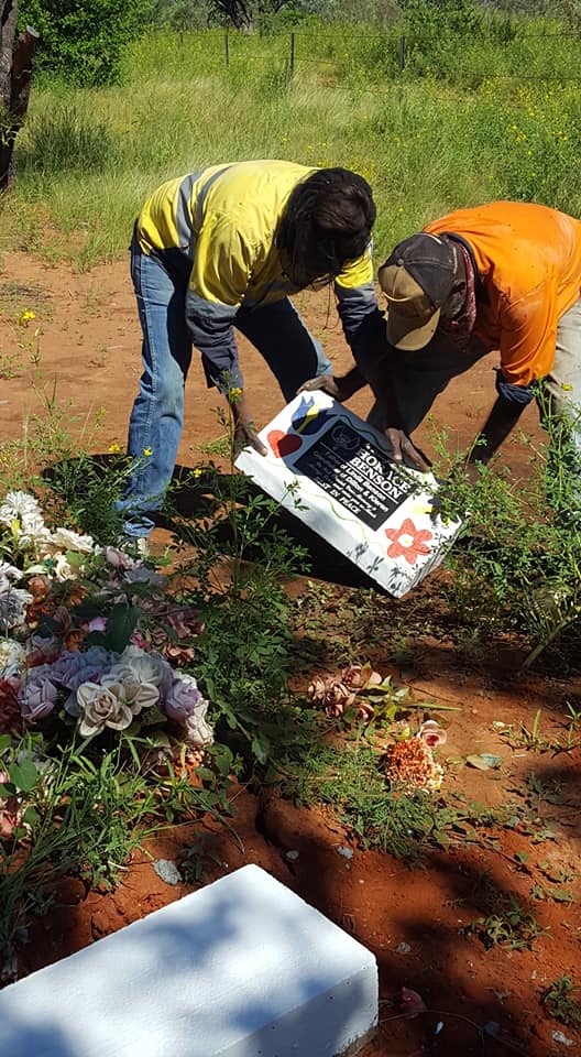 Two people in work clothes tending a grave, placing a decorative plaque near flowers.