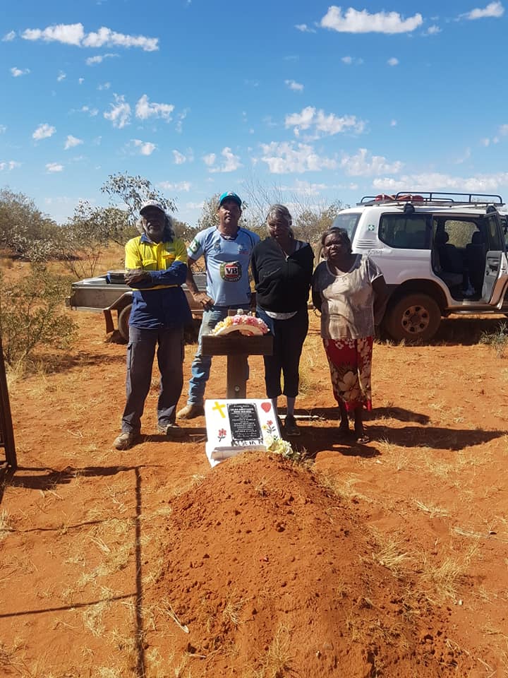 Four people stand near a freshly dug grave in a dry, rural area. A car and a table with items are visible.