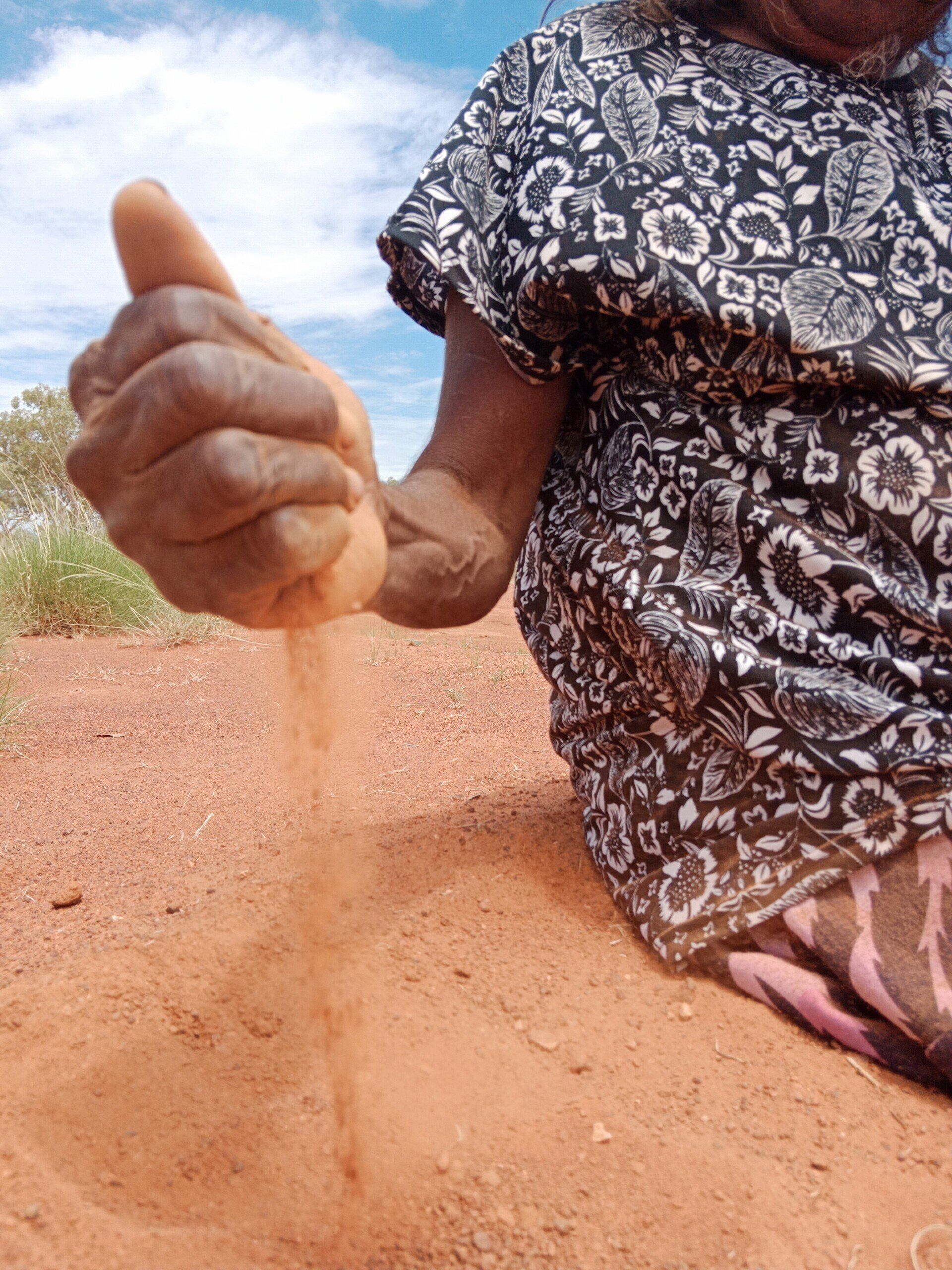 An Indigenous person's hand releasing red sand. The person sits on the ground in a sunny, desert-like environment.