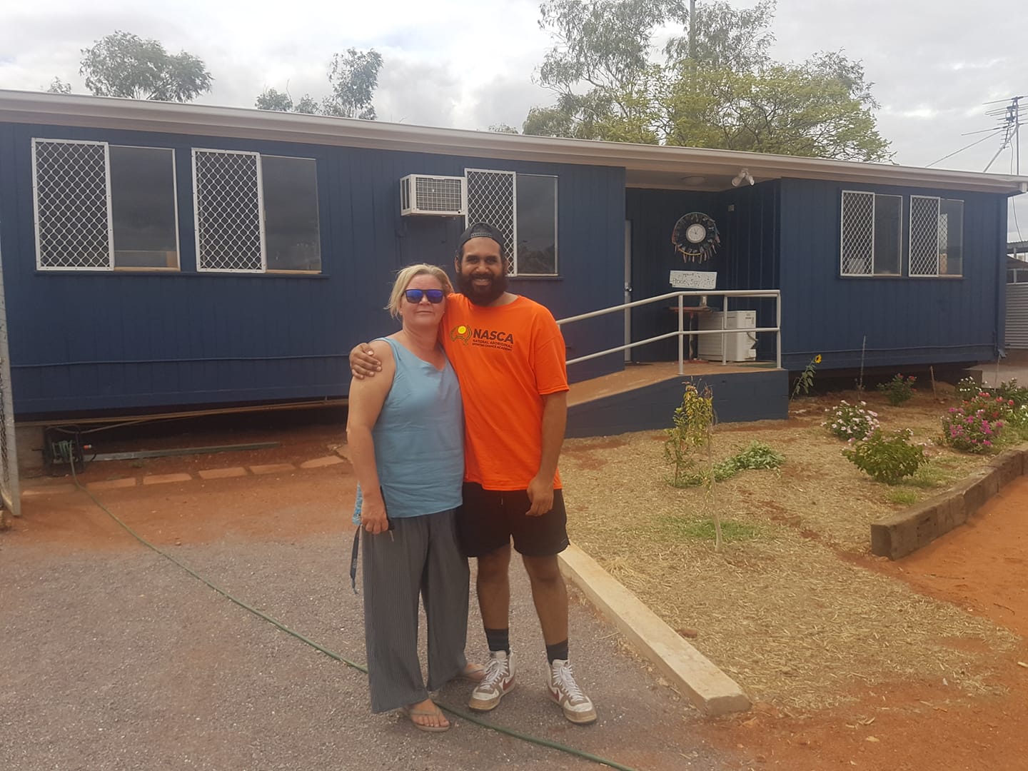 Two people posing in front of a blue building with windows.