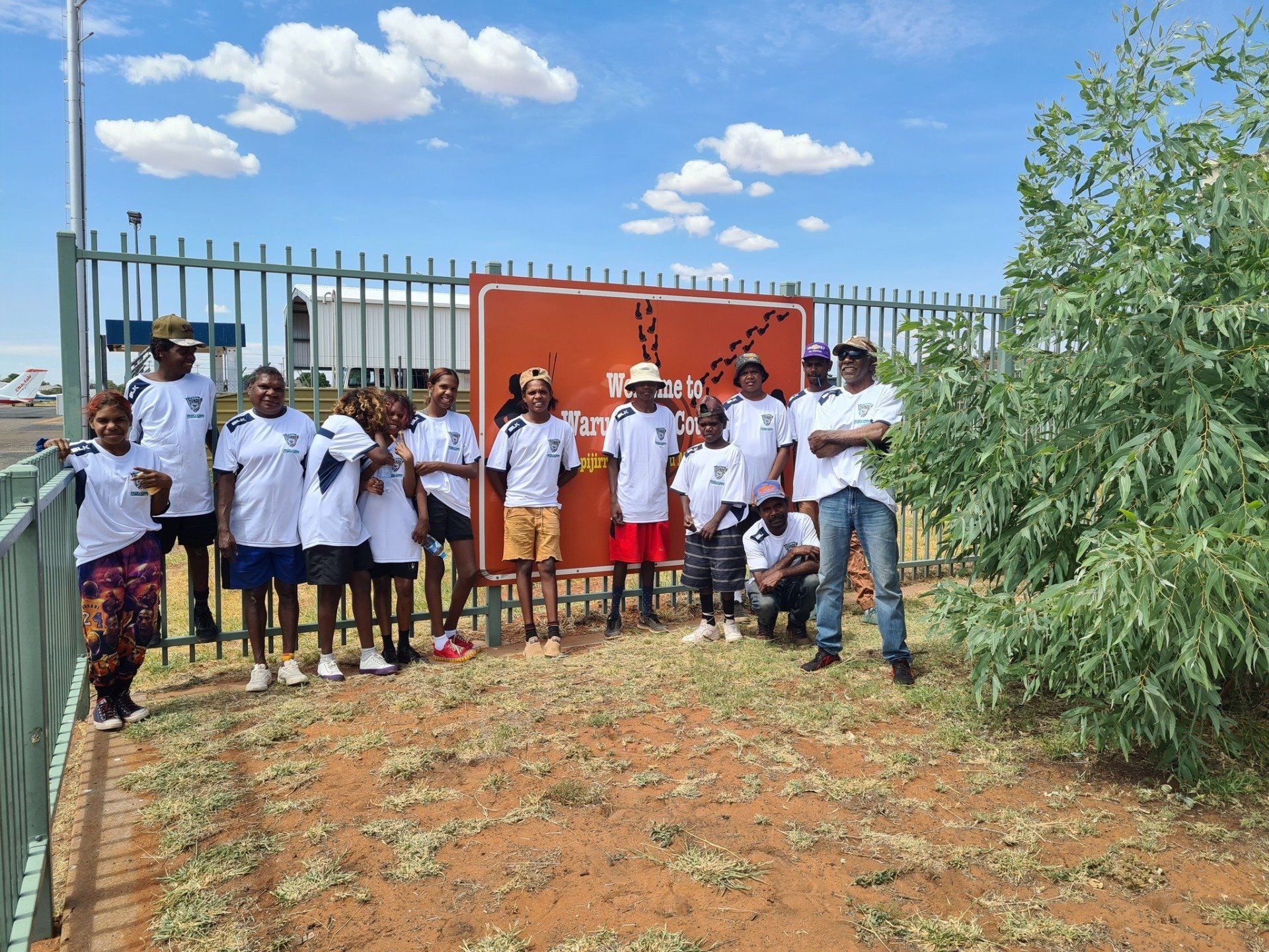 A group of people in white shirts stand in front of a fence and sign outdoors under a blue sky. They are smiling.