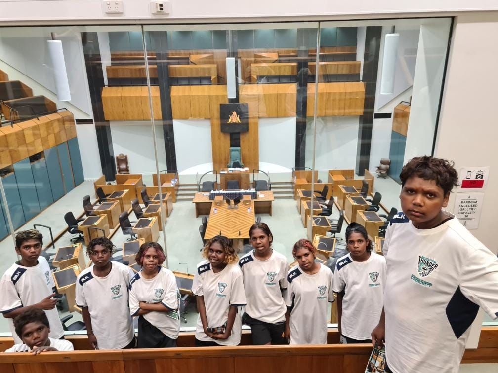 A group of people in matching white shirts pose inside a parliamentary chamber, looking at the camera.