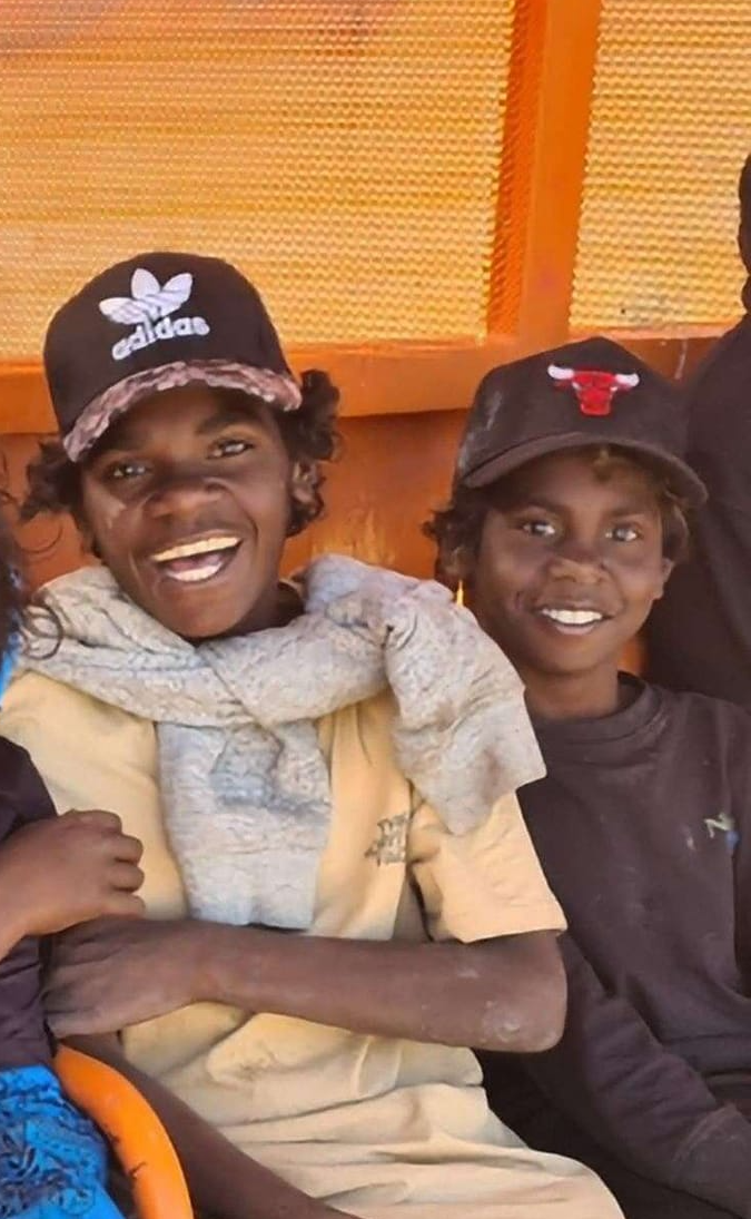Two young Indigenous people smiling, one wearing an Adidas cap and scarf.