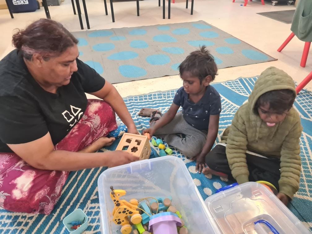 A woman and two children sit on a blue mat playing with toys, including a shape sorter. 
