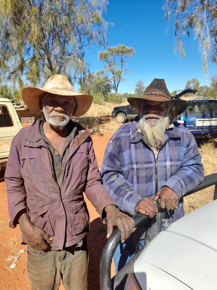 Two older Indigenous men in hats and jackets, outdoors in a sunny, dusty setting. One leans on a car.