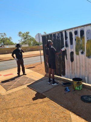 Two people painting an outdoor wall. One paints while the other watches, both in casual clothing under a bright sun.