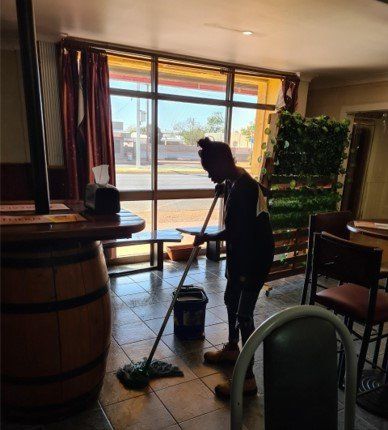 A person mops the floor of a restaurant near a window. Sunlight streams in; a wooden barrel and vertical garden are visible.