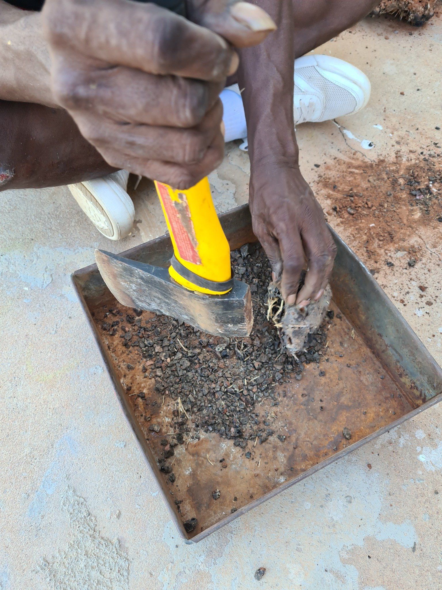 Person using a hand tool to sift through dark material in a metal tray, outdoors.