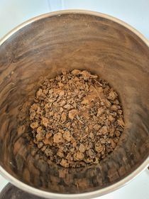 A metal container holds a layer of brown, chunky, dried material, possibly soil or plant matter.