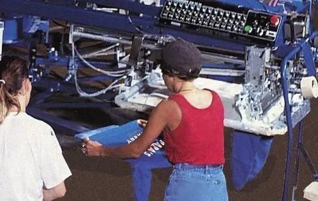 Workers operating a large blue printing press, feeding material into the machine.