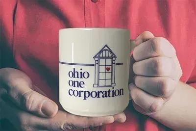 Hands holding a white mug with an Ohio One Corporation logo and house graphic.